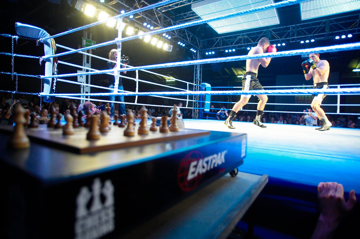 Two chessboxers trading punches in a lit ring, with the chessboard waiting on its trolley in the foreground.