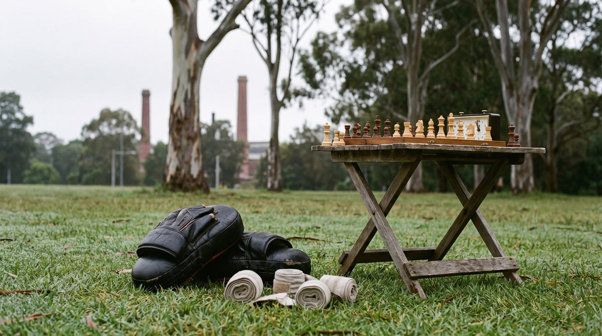 Boxing pads and hand wraps on the grass beside a folding table with a chess set, Sydney Park brickworks chimneys behind.